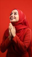 Arabic woman wearing scarf is praying and smiling on red background photo