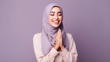 Arabic woman wearing scarf is praying and smiling on purple background photo