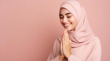 Arabic woman wearing scarf is praying and smiling on pink background photo