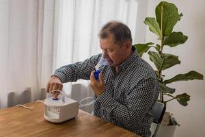 mayor hombre sentado en un mesa y utilizando un nebulizador niebla a hogar foto