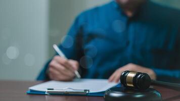 A lawyer man is writing on a piece of paper in front of a gavel. The gavel is on a desk and the man is sitting at a table photo