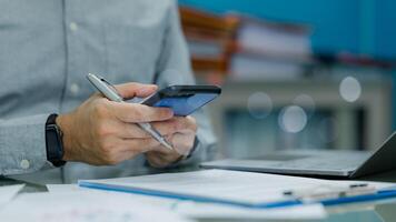 A businessman or businessperson is using a cell phone while sitting at a desk with a laptop and a stack of papers photo