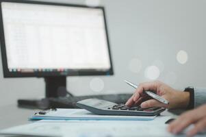 un persona es utilizando un calculadora en un escritorio con un computadora monitor en el antecedentes. el persona es enfocado en el calculadora y él es trabajando en un tarea. concepto de concentración y productividad foto