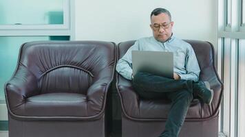 A businessman is sitting on a leather chair with a laptop in front of him. He is wearing glasses and he is focused on his work. Concept of productivity and concentration photo