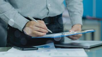 Businessman is sign accounting on a clipboard with a pen in office. The clipboard is blue and has a white border. ceo man is wearing a gray shirt and black pants photo