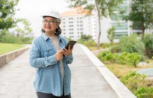 Happy elderly Asian professional engineer wears a helmet and checks the blueprint on a tablet on the construction site after the contractor and architect finish renovation the house and building photo