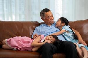 Happy Asian senior grandfather sits on a couch with his granddaughter and plays with his granddaughter in the living room at home, The Concept of family having fun in their house photo