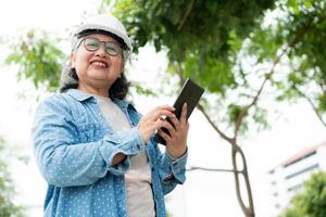 Happy elderly Asian professional engineer wears a helmet and checks the blueprint on a tablet on the construction site after the contractor and architect finish renovation the house and building photo