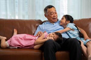 contento asiático mayor abuelo se sienta en un sofá con su nieta y obras de teatro con su nieta en el vivo habitación a hogar, el concepto de familia teniendo divertido en su casa foto