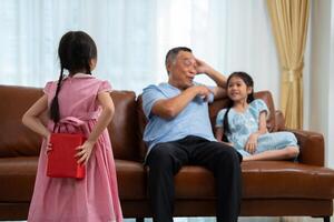 pequeño niña ocultación presente caja detrás espalda mientras haciendo sorpresa mayor abuelos, alegre mayor hombre sonriente y gesticulando mientras recibe un sorpresa regalo desde nieta para celebracion foto