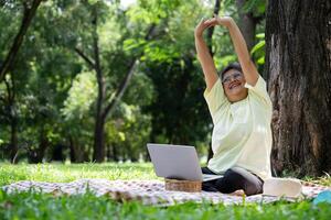 Happy Asian senior woman using laptop for working online outside office. Elderly retirement using laptop for learning new skill and freelance working in park, senior lifestyle with technology photo