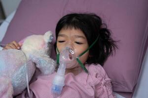 un pequeño asiático niña tiene un oxígeno máscara y respiración mediante un nebulizador a el hospital. concepto de bronquitis, respiratorio y médico tratamiento, inhalando medicamento, neblina, desde un nebulizador. foto