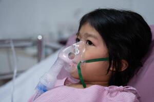 un pequeño asiático niña tiene un oxígeno máscara y respiración mediante un nebulizador a el hospital. concepto de bronquitis, respiratorio y médico tratamiento, inhalando medicamento, neblina, desde un nebulizador. foto