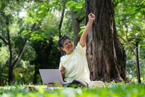 Happy Asian senior woman using laptop for working online outside office. Elderly retirement using laptop for learning new skill and freelance working in park, senior lifestyle with technology photo