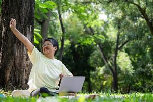 Happy Asian senior woman using laptop for working online outside office. Elderly retirement using laptop for learning new skill and freelance working in park, senior lifestyle with technology photo