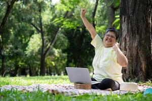 Happy Asian senior woman using laptop for working online outside office. Elderly retirement using laptop for learning new skill and freelance working in park, senior lifestyle with technology photo