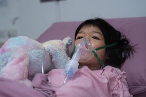 un pequeño asiático niña tiene un oxígeno máscara y respiración mediante un nebulizador a el hospital. concepto de bronquitis, respiratorio y médico tratamiento, inhalando medicamento, neblina, desde un nebulizador. foto