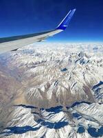 Captivating aerial shot of snow-capped mountains beneath a plane's wing, showcasing the grandeur of nature's peaks against a clear blue sky photo