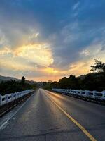 A tranquil highway leads towards a stunning sunset, flanked by lush greenery and radiant skies at dusk. photo