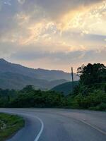 A tranquil highway leads towards a stunning sunset, flanked by lush greenery and radiant skies at dusk. photo