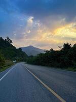 A tranquil highway leads towards a stunning sunset, flanked by lush greenery and radiant skies at dusk. photo