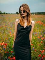 Beautiful young girl in a black evening dress and sunglasses posing against a poppy field on a cloudy summer day. Portrait of a female model outdoors. Rainy weather. Gray clouds. photo