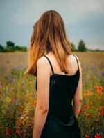 Beautiful young girl in black evening gown, standing back, posing against poppy field on cloudy summer day. Portrait of female model outdoors, rear. Rainy weather. Gray clouds. photo