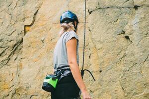 joven atlético mujer en equipo en pie en frente de Roca rock al aire libre y consiguiendo Listo a trepar. formación zona para al aire libre actividades. extremo deporte. posterior ver foto