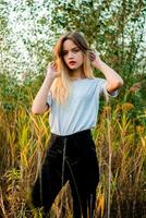 hermosa joven niña vistiendo blanco gris camiseta y negro pantalones posando en contra alto verde y amarillo césped en temprano calentar otoño. al aire libre retrato de hermosa hembra modelo foto