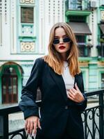 A woman in sunglasses, a white shirt and a black jacket stands confidently on the street. photo