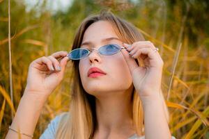 Beautiful young girl wearing blank gray t-shirt and black jeans in blue glasses posing against high green and yellow grass in early warm autumn. Outdoor portrait of beautiful female model. photo