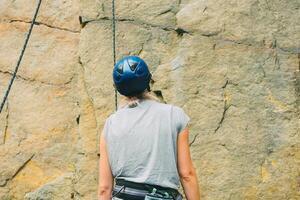 Young athletic woman in equipment standing in front of stone rock outdoors and getting ready to climb. Training area for outdoor activities. Extreme sport. Rear view photo