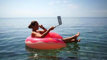 mujer trabajos en ordenador portátil en mar. persona de libre dedicación, rubio mujer en gafas de sol flotante en un inflable grande rosado rosquilla con un ordenador portátil en el mar a puesta de sol. personas verano vacaciones descanso estilo de vida concepto. video