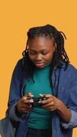 Vertical Euphoric african american woman playing intense gaming console game, celebrating win, studio background. Ecstatic young girl using controller, excited after being victorious in videogame, camera A video