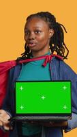 Vertical Portrait of jolly teenager acting as superhero presenting green screen laptop, isolated over studio background. Cheerful BIPOC person posing as hero showing mockup notebook features, camera A video