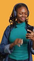 verticale africano americano adolescente parla con amici durante video conferenza utilizzando Telefono e cuffia, studio sfondo. giovane bipoc ragazza chat con compagni durante Internet video chiamata su cellulare, telecamera un'