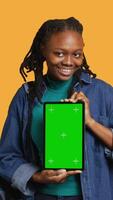 Vertical Smiling african american woman presenting tablet with green screen display, isolated over studio background. Cheerful BIPOC person creating promotion with chroma key device, camera A video