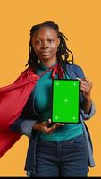 Vertical Portrait of joyful woman portraying superhero showcasing green screen tablet, giving positive feedback, studio background. Radiant person doing hero cosplay recommending mockup device, camera B video