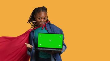 Portrait of BIPOC woman portraying superhero with cape presenting isolated screen laptop, studio background. African american young girl posing as hero talking, showing chroma key notebook, camera A video