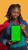 Vertical Portrait of joyful woman portraying superhero showcasing green screen tablet, giving positive feedback, studio background. Radiant person doing hero cosplay recommending mockup device, camera A video