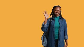 retrato de alegre niña participación mochila, ondulación mano, gesticulando, aislado terminado amarillo estudio antecedentes. alegre africano americano estudiante con colegio mochila haciendo saludo mano gesto, cámara si video