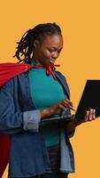 Vertical Happy african american young girl wearing superhero for Halloween working on laptop, studio background. Jolly teenager dressed as comic book hero typing on notebook keyboard, camera B video