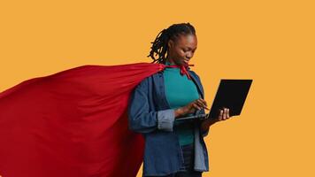 Happy african american young girl wearing superhero for Halloween working on laptop, studio background. Jolly teenager dressed as comic book hero typing on notebook keyboard, camera B video