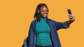 Cheerful woman with headphones using smartphone to take selfies and post them on social media. Happy person taking photos using phone selfie camera, isolated over studio background video