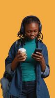 Vertical Woman scrolling on smartphone, using phone to check social media feed, drinking coffee. Girl browsing internet websites, enjoying beverage, listening music, isolated over studio background, camera B video