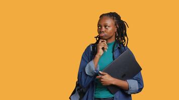 Pensive woman carrying laptop, doing thinking gesture, memorizing information for school exam, studio background. Student holding notebook, remembering before university assessment, camera A video