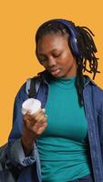 Vertical Young girl with backpack and headphones yawning, feeling tired, drinking coffee to wake up. Sleepy woman using caffeinated beverage to gain energy, lacking sleep, studio background, camera A video