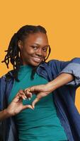 Vertical Portrait of friendly smiling african american woman doing heart symbol shape gesture with hands, being affectionate. Cheerful nurturing person showing love gesturing, studio background, camera A video