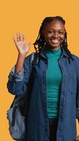 Vertical Portrait of chipper girl holding backpack, waving hand, gesturing, isolated over yellow studio background. Jolly african american student with school rucksack doing greeting hand gesture, camera B video