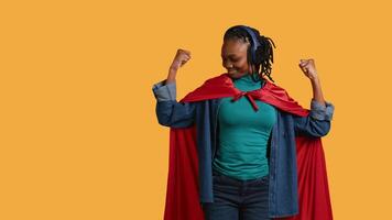 African american superhero with red cape flexing muscles, isolated over studio background. Woman wearing cloak posing as hero in costume kissing biceps, showing strength, camera B video